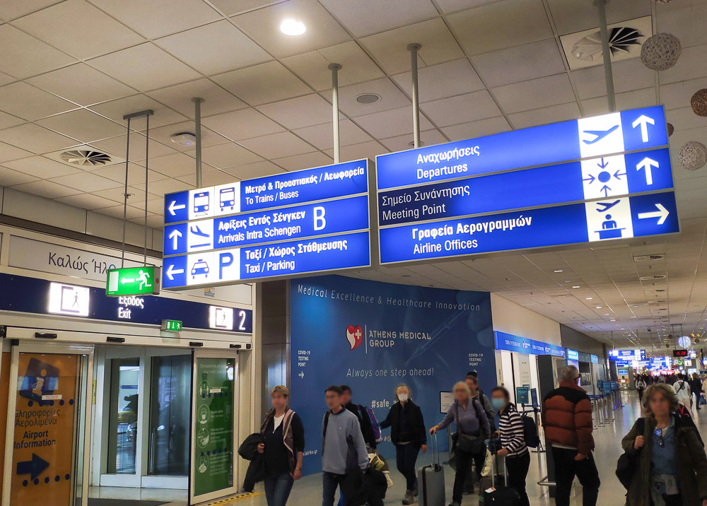 The Athens airport arrivals corridor with blue coloured signage with white text showing the way to the public bus stand.